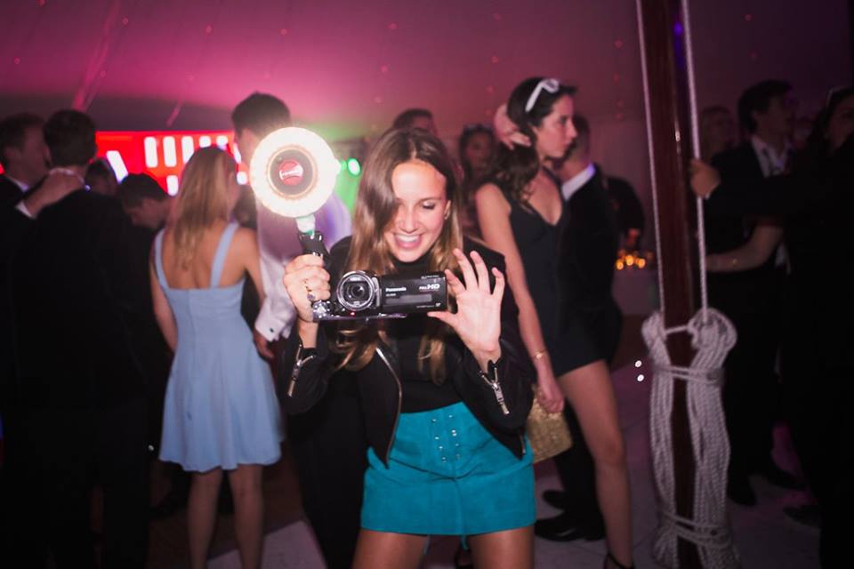Girl at a party in blue skirt and black leather jacket holding a video camera with flash