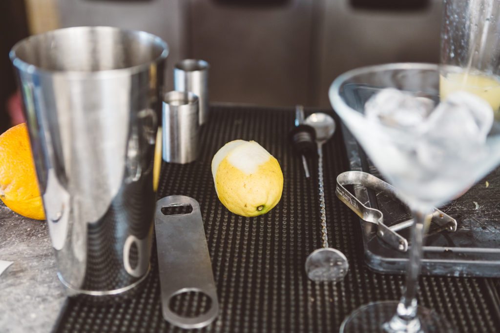 Cocktail making equipment with a martini glass with ice and a silver metal shaker, a semi peeled lemon and a silver bottle opener