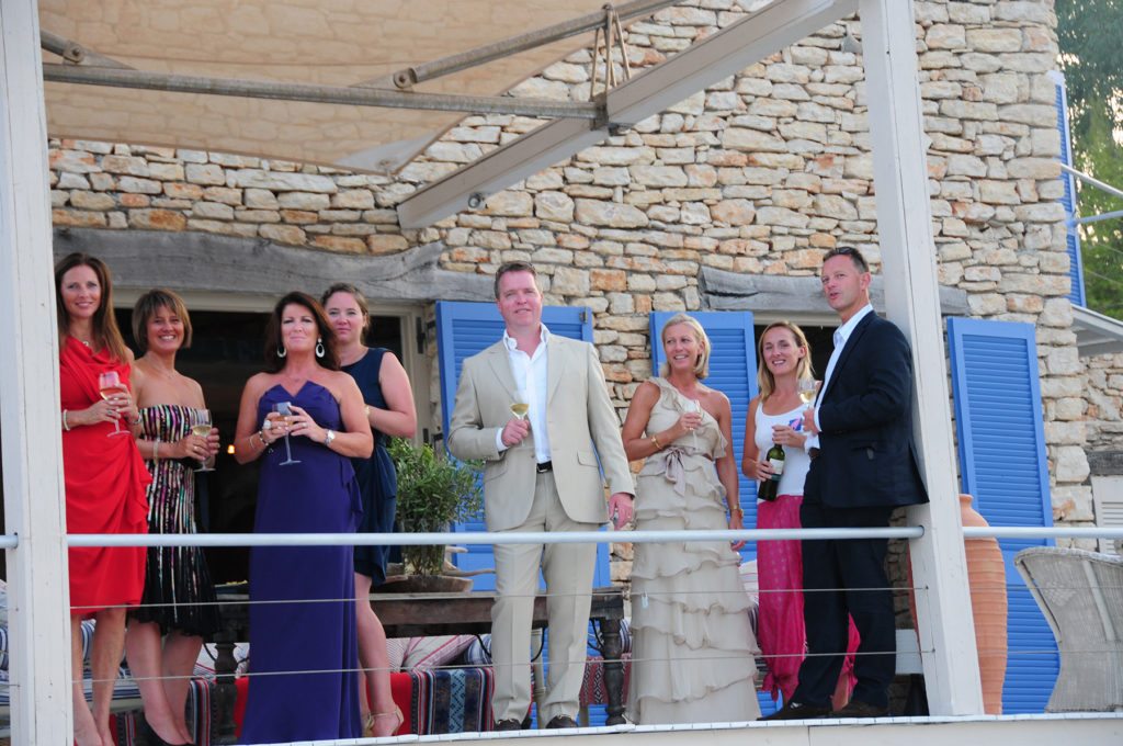Guests wearing glamorous colourful dresses on the terrace of a luxury villa having a drinks party 